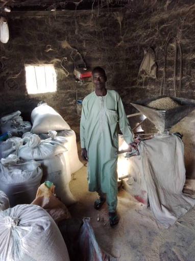 A Nigerian man posing next to a diesel rice mill in a room full of bags of rice.