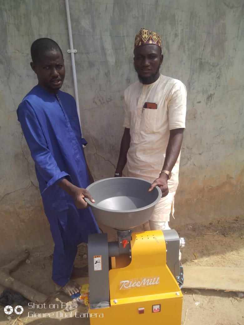 Two Nigerian men standing next to a yellow rice mill.